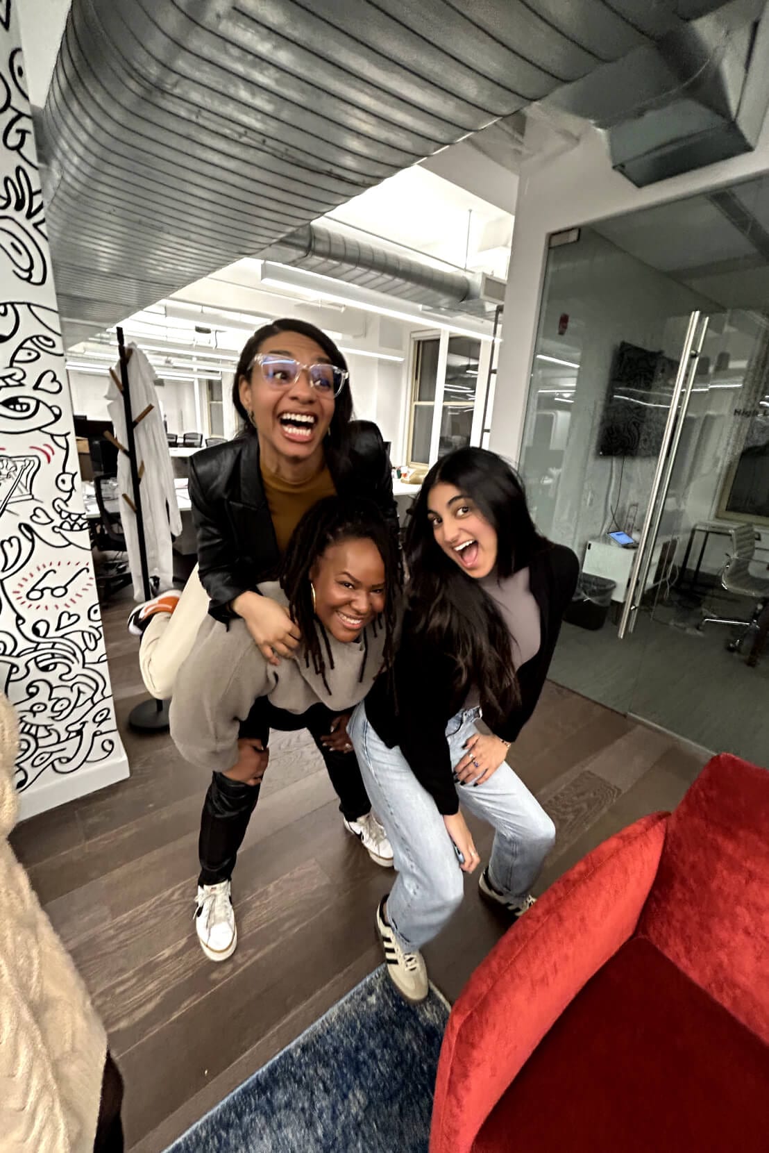Three women are having fun in an office setting. One woman is giving a piggyback ride to another woman, while a third woman poses beside them